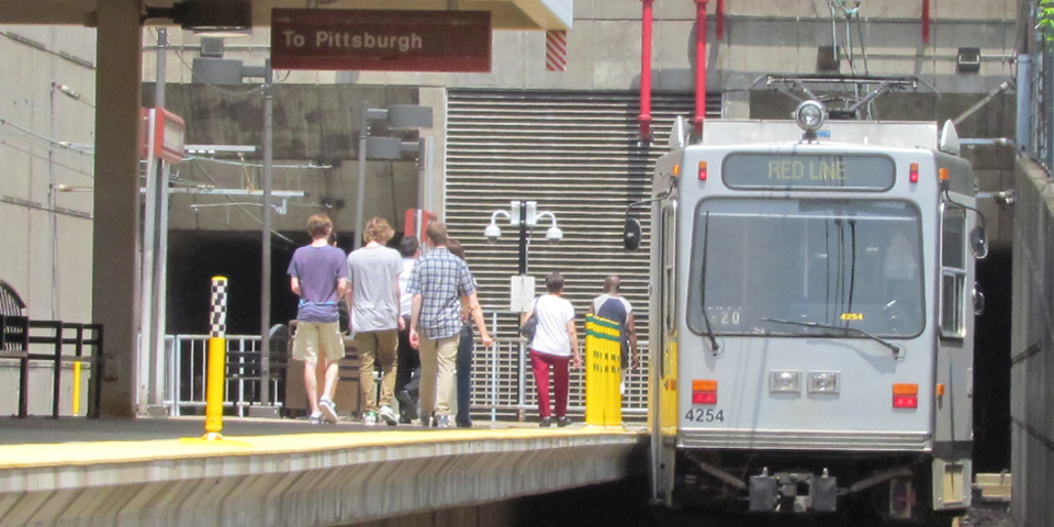 The T pulling into the Mt. Lebanon station with a group of people walking towards it to get on. The sign at the stop reads "To Pittsburgh" and on the trolley it reads "Red Line."