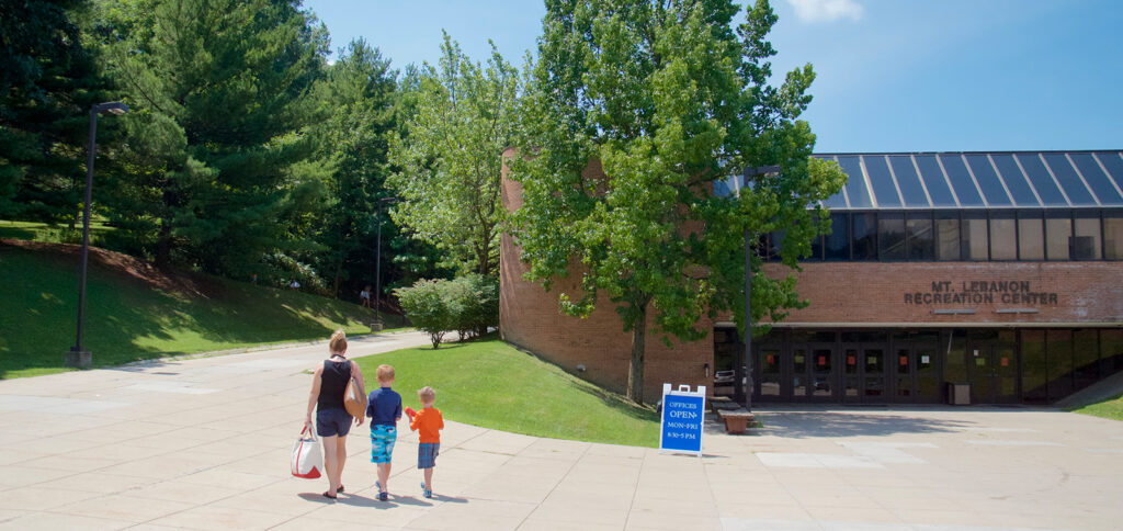 The front of the Mt. Lebanon Recreation Center with a mom and two young children walking toward the pool path.